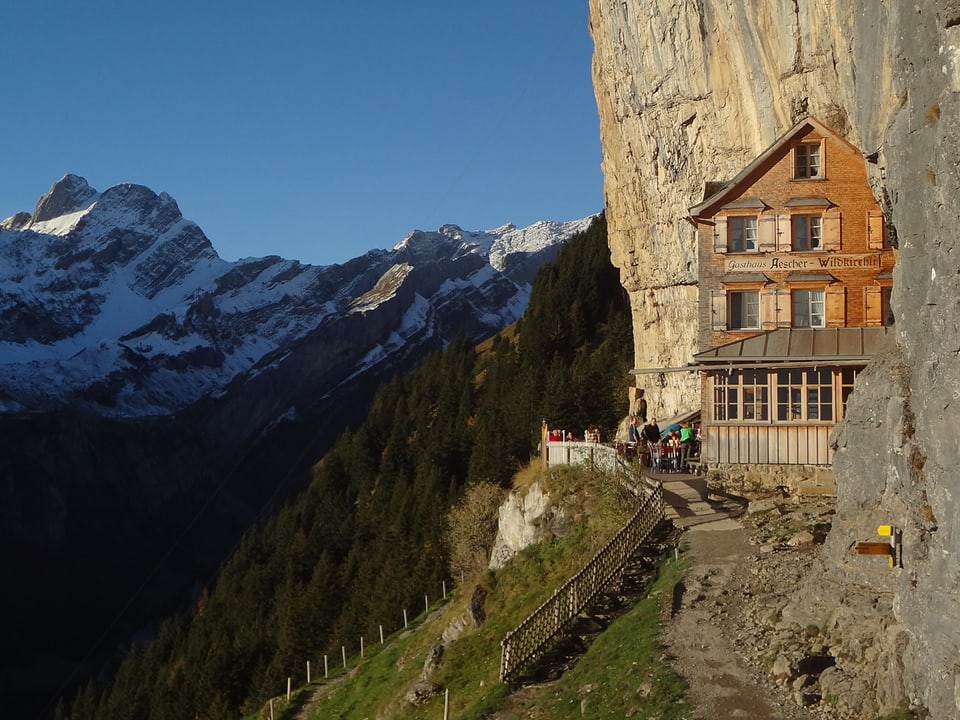 Ein Haus klebt an einer Felswand, die verschneiten Berge im Hintergrund.