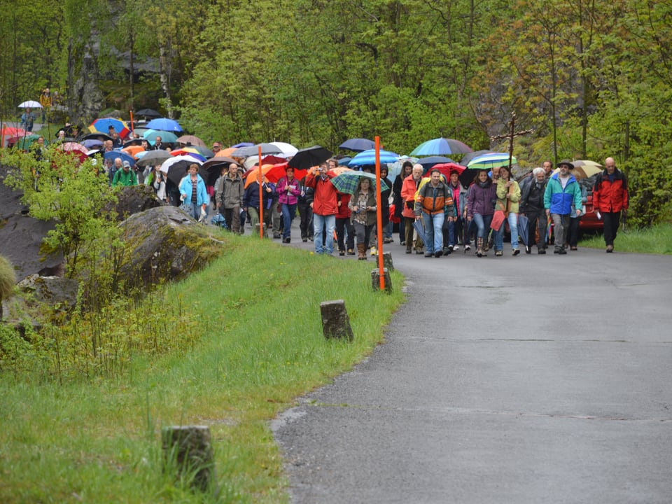 Eine grosse Gruppe Menschen in farbiger Regenkleidung wandert durchs Tal. Zuvorderst ein Kruzifix. 