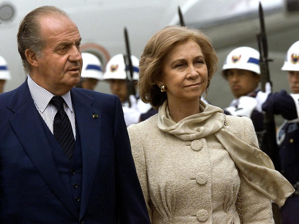 König Juan Carlos in dunkel blauem Anzug mit Königin Sofia in heller Jacke laufen neben Soldaten.