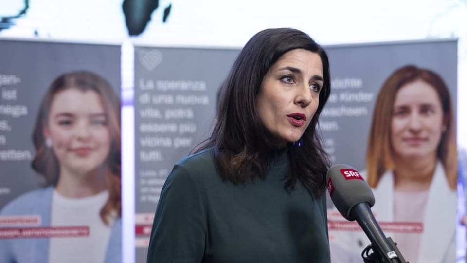 Frau mit schulterlangen schwarzen Haaren. Sie trägt einen dunkelgrünen Pulli und steht vor Abstimmungsplakaten am Mik.