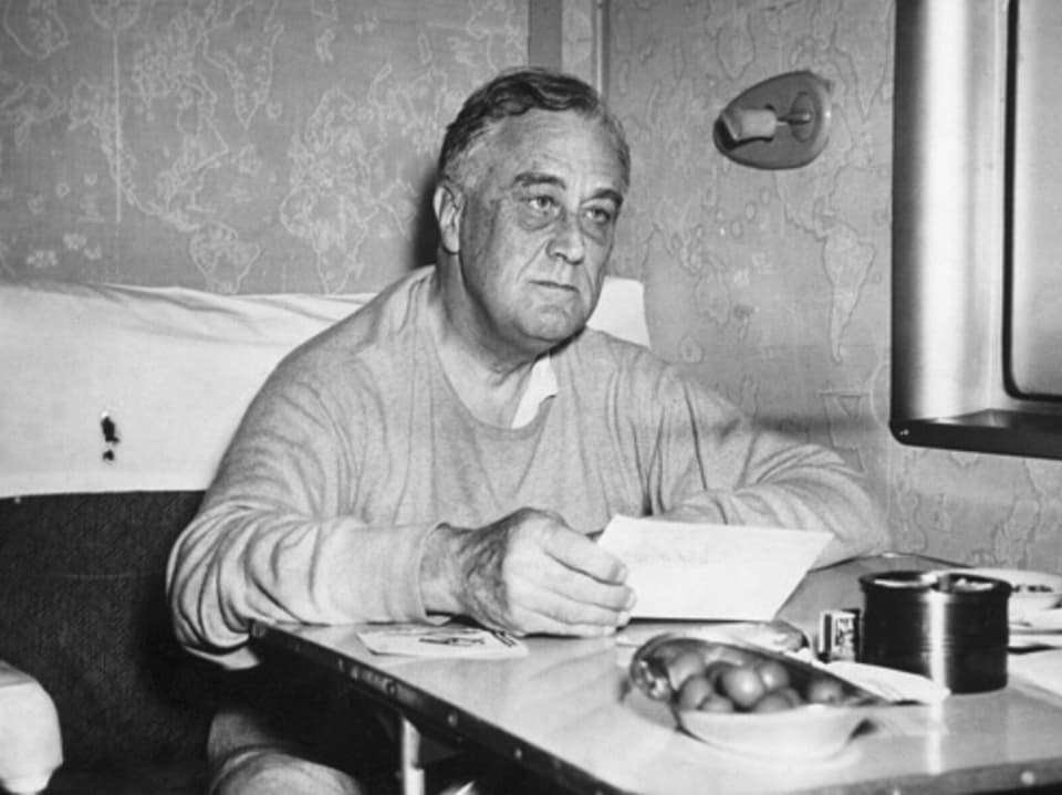 Krank aussehender Franklin D. Roosevelt sitzt an einem Tisch und hat ein Blatt Papier in der Hand.