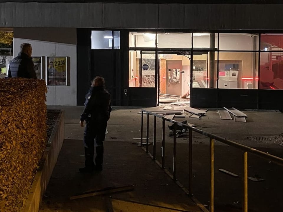 Zwei Mitarbeitende der Polizei stehen vor der Bankfiliale mit dem gesprengten Automaten.