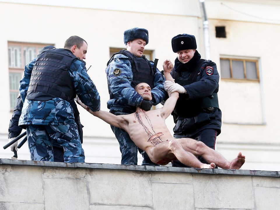 Drei Polizisten halten einen nackten Mann fest. Auf seinem Oberkörper hat es Blutspuren.