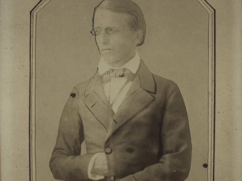 Schwarz-Weiss-Foto eines Mannes, der seine rechte Hand auf der Höhe seines Bauches in die Öffnung seines Mantels steckt. 