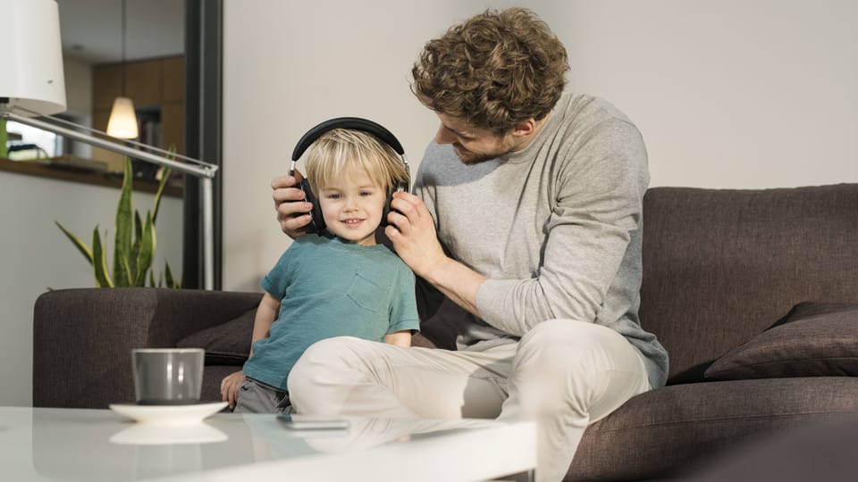 Vater zieht seinem Kind Kopfhörer an auf einem Sofa.