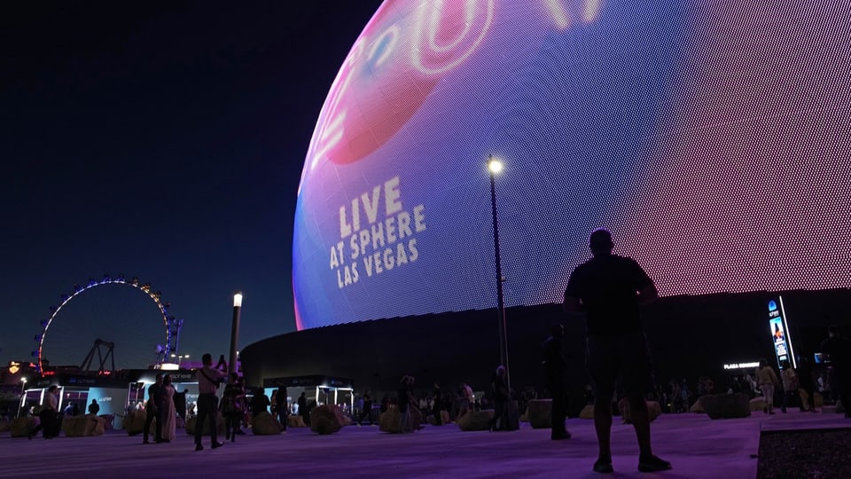 Besucherinnen und Besucher stehen vor der leuchtenden Kuppel der Mega-Eventhalle Sphere.