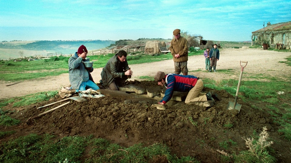Vier Männer buddeln mit Schaufeln eine Grube, zwei Kinder stehen im Hintergrund und beobachten sie.