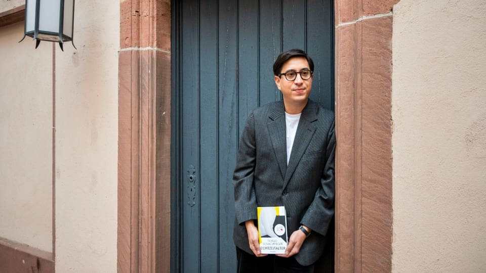 Ein junger Mann mit runder Brille steht in einem Hauseingang. Er hält ein Buch in beiden Händen.