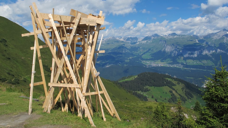 Ein Holzgerüst, das an einen Turm erinnert inmitten einer alpinen Sommerlandschaft.