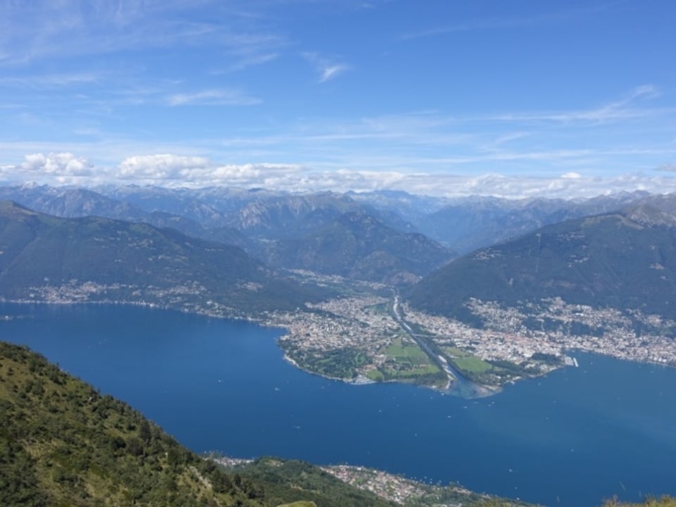Blick vom Monte Gambarogno auf den Lago Maggiore und das Maggiadelta. Dahinter Quellwolken über den Bergen des Maggiatals und den anschliessenden Walliser Bergen.