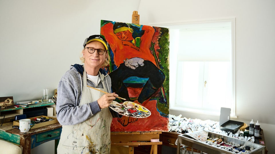 Mann mit kurzen blonden Haaren und Kappe steht vor einer Staffelei mit einem Gemälde. Er trägt Malerkittel und Palette.