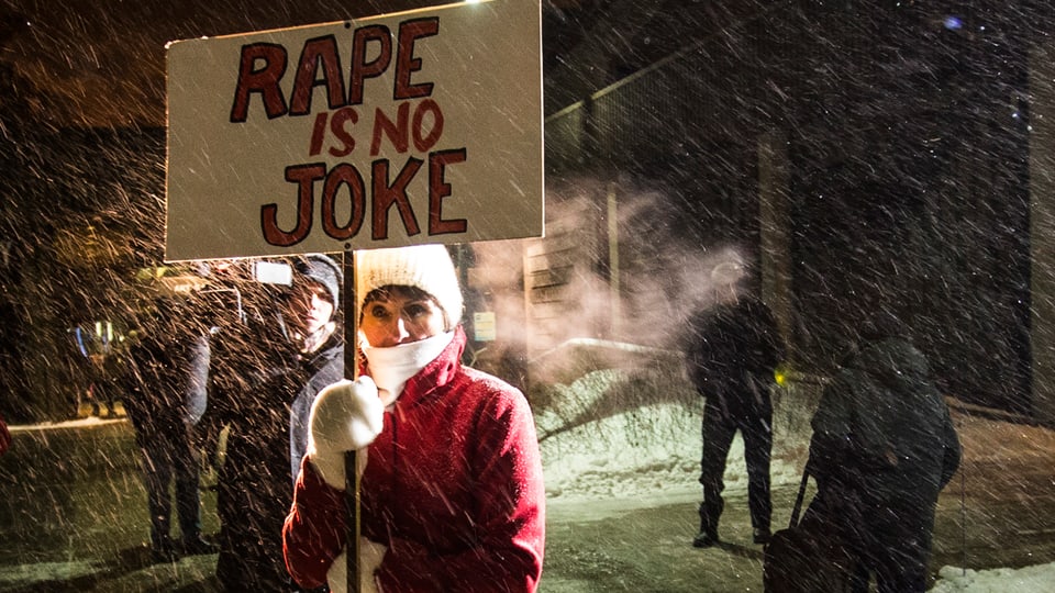 Eine Frau in einem roten Mantel hält ein weisses Transparent in der Hand. Darauf ist geschrieben: «Rape is no Joke».