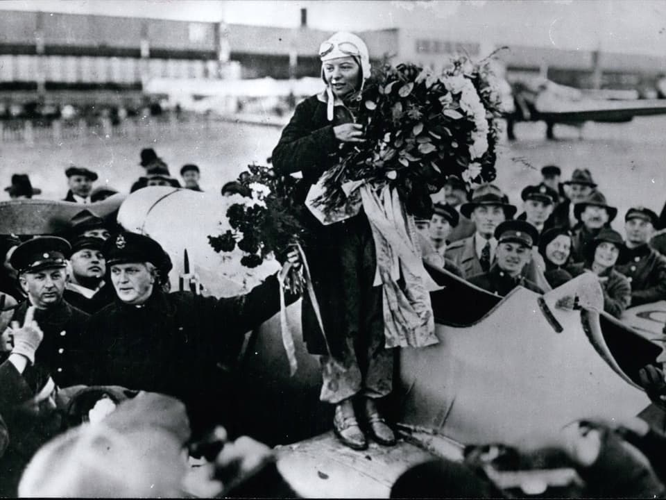 Elly Beinhorn steht mit einem Blumenstrauss auf einem Flugzeugflügel und wird nach ihrem Flug um die Welt bejubelt.