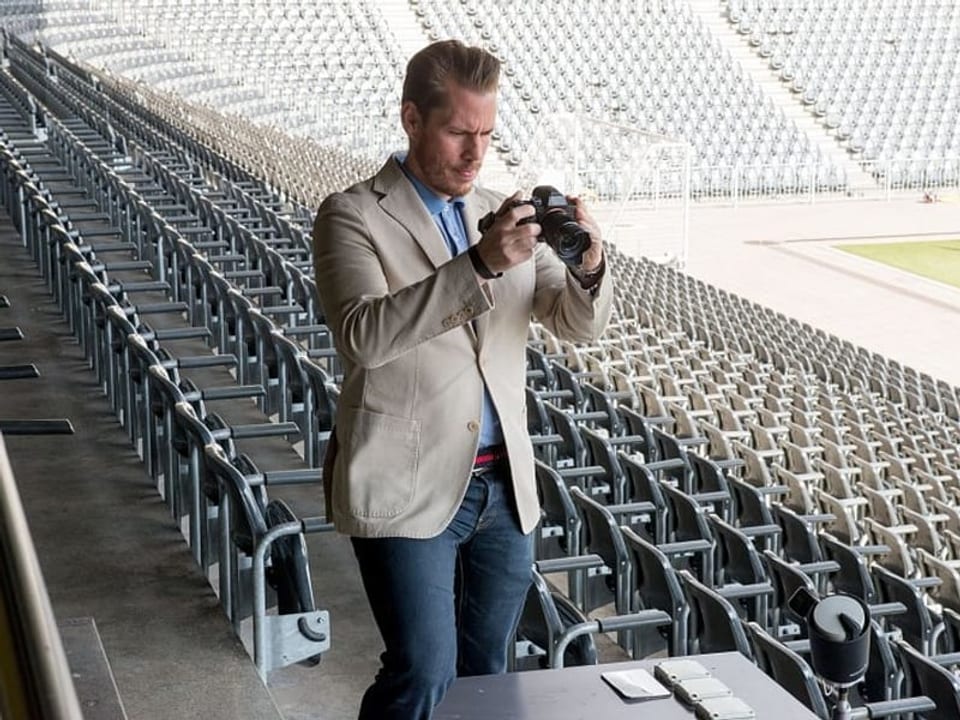 Ein Mann mit Anzug und Hemd und einer Kamera in der Hand steht in einem leeren Fussballstadion. 