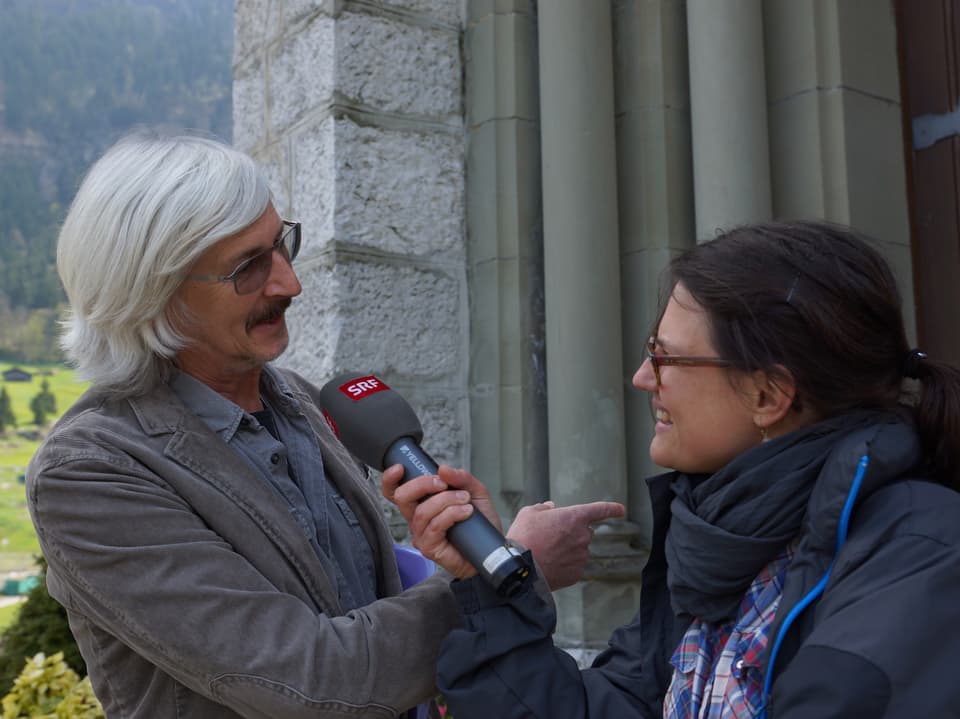 Ein Mann und eine Frau unterhalten sich übers Mikrofon vor einem Kirchenportal.
