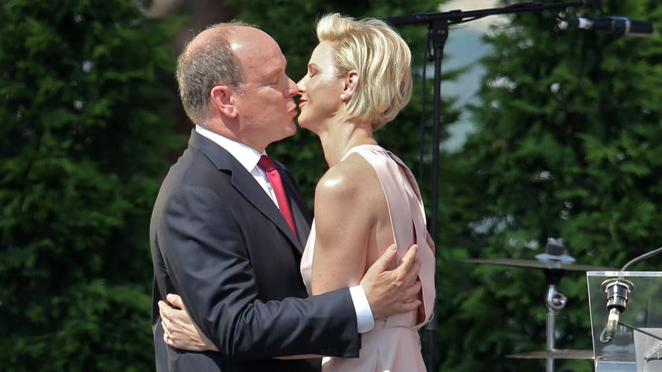 Fürst Albert steht im Anzug links und küsst seine Frau Charlène, die recht steht und ein freizügiges, rosarotes Kleid trägt. 