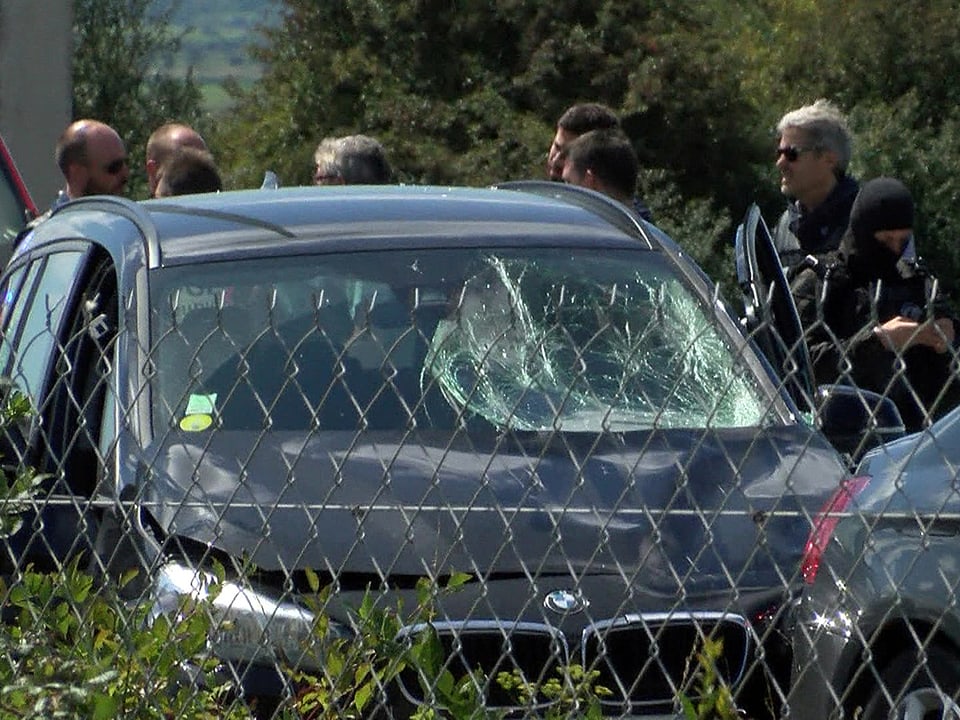 Ein schwarzer BMW mit kaputter Scheibe. Mehrere Sicherheitskräfte stehen daneben.