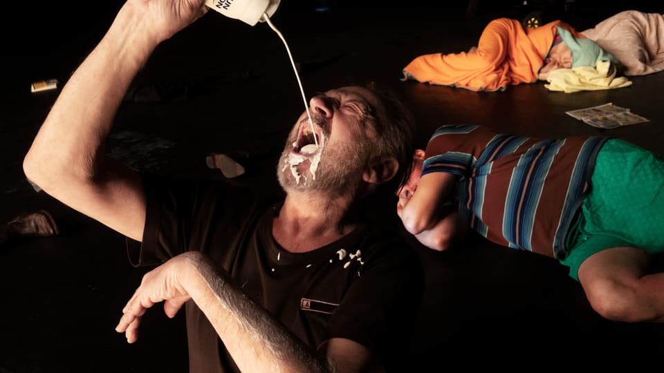 A man pours milk into his mouth.  He's visibly making a mess.  behind him there are people on the stage.