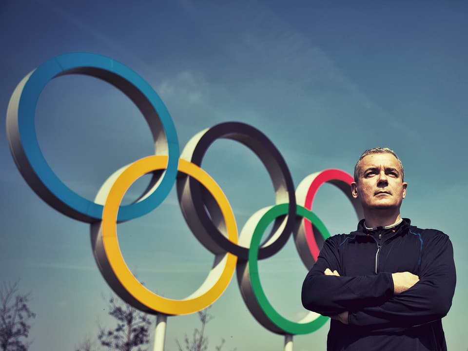 Der Schriftsteller Ilija Trojanow steht mit verschränkten Armen neben den olympischen Ringen.