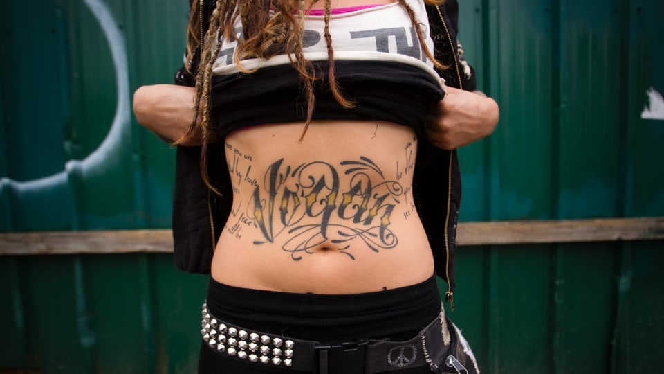 Frau hält Tshirt hoch. Auf dem Bauch hat sie ein Tattoo. Es steht vegan drauf.