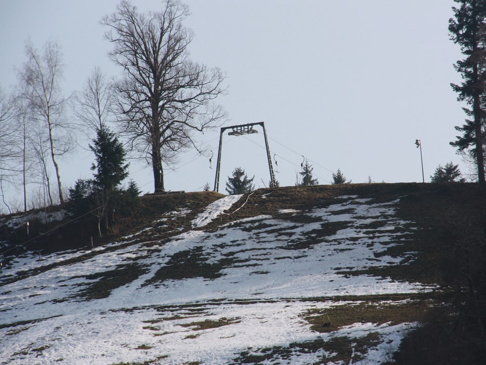 Blick auf einen Hang mit ein paar weissen Schneeflecken und den Pfosten eines Skilifts.