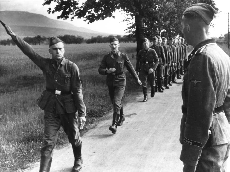 Soldaten marschieren hintereinander auf einem Feldweg. Der Forderste hebt den Arm zum Hitler-Gruss.