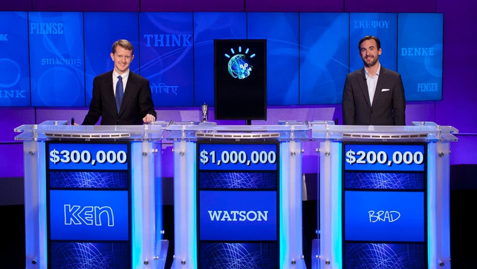 Zwei Männer und ein Computer stehen hinter den Kandidatenpulten der US-Quizshow Jeopardy!