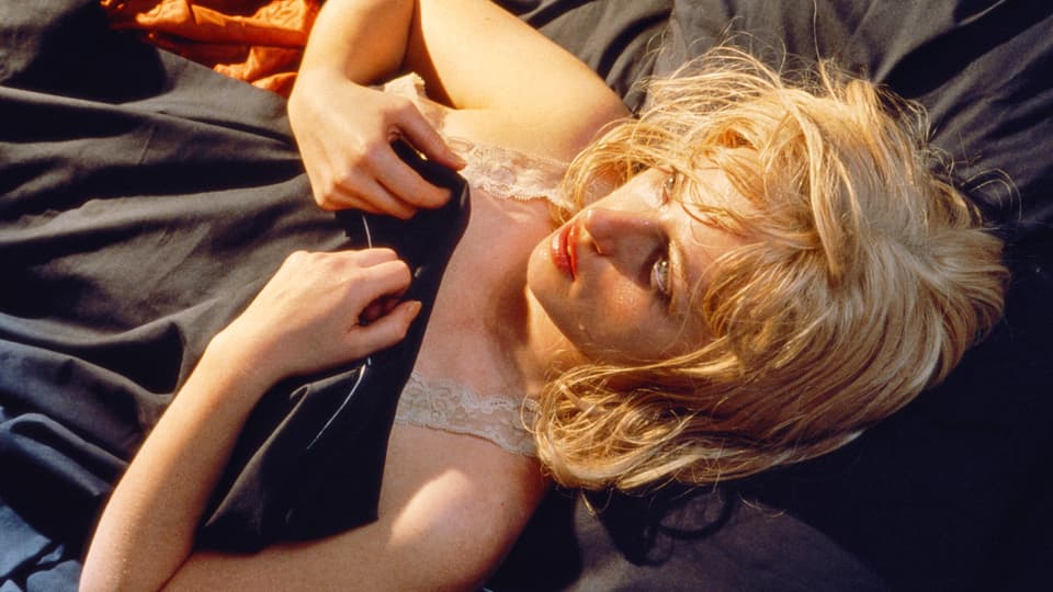 Eine blonde Frau mit geschminkten Lippen und Augen liegt unter einer schwarzen Decke.