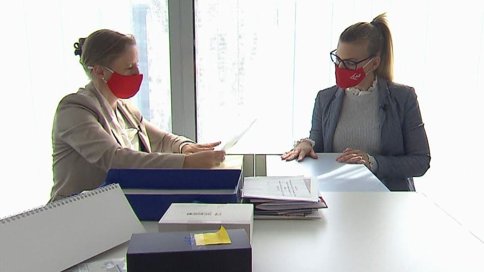 Zwei Frauen in Anzügen und mit roten Masken sitzen an einem Tisch. Auf dem Tisch liegen Papier und Ordner.