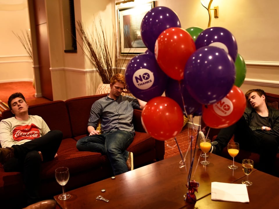 Drei Männer schlafen auf dem Sofa, auf dem Tisch sind Nein-Ballone der Referendums-Gegner