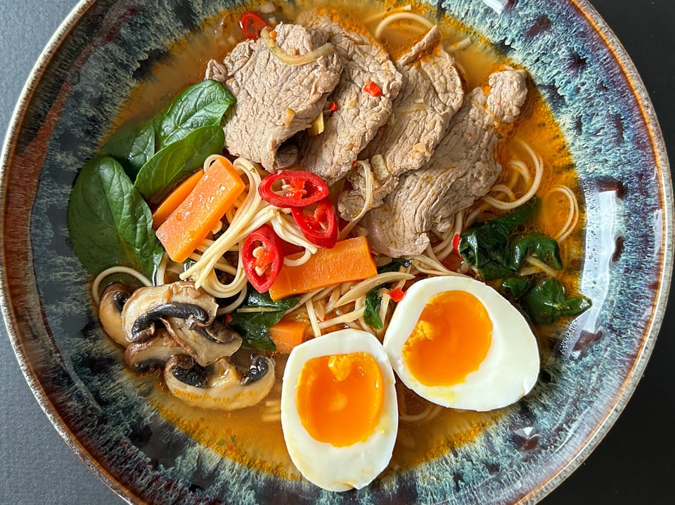 Asiatische Nudelsuppe mit Rindfleisch, Gemüse, Pilzen und halbierten Eiern auf einem bunten Teller.