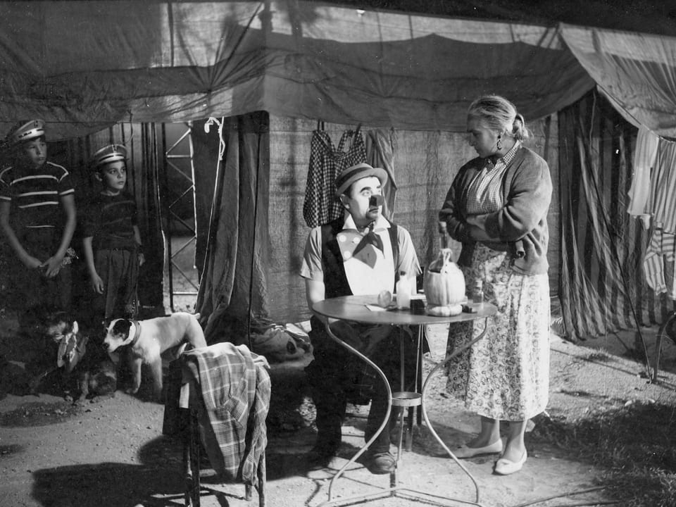 Ein Mann mit einer Clownsnase sitzt an einem Tisch. Vor ihm steht Wein. Eine Frau steht mit verschränkten Armen neben ihm. Im Hintergrund ist ein grosses Zelt zu sehen, zwei jungen stehen mit ihrem Hund daneben und blicken auf die beiden.