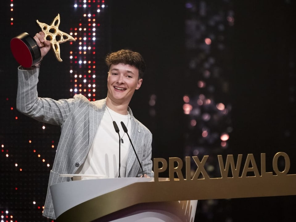 Junger Mann hebt einen Preis auf einer Bühne mit Schriftzug 'PRIX WALO' in die Höhe.