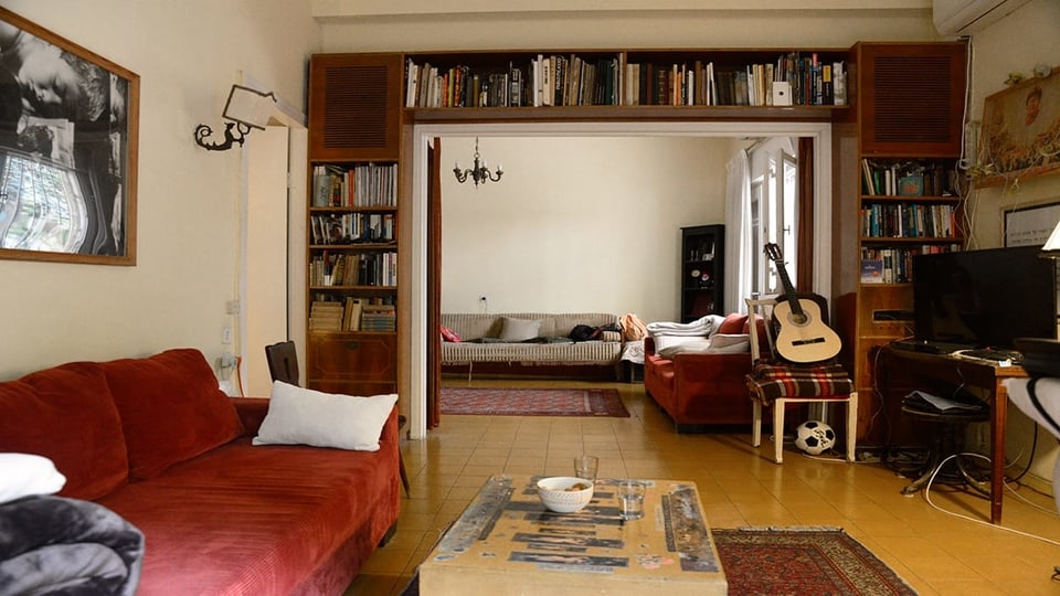 Blick in ein gemütliches Wohnzimmer. Ein rotes Sofa, viele Bücher, ein Stuhl mit einer Gitarre drauf.