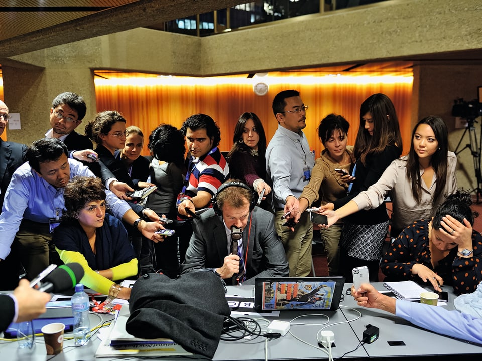 Eine Horde Journalisten mit Mikrofonen lauert um einen Tisch mit Laptop.