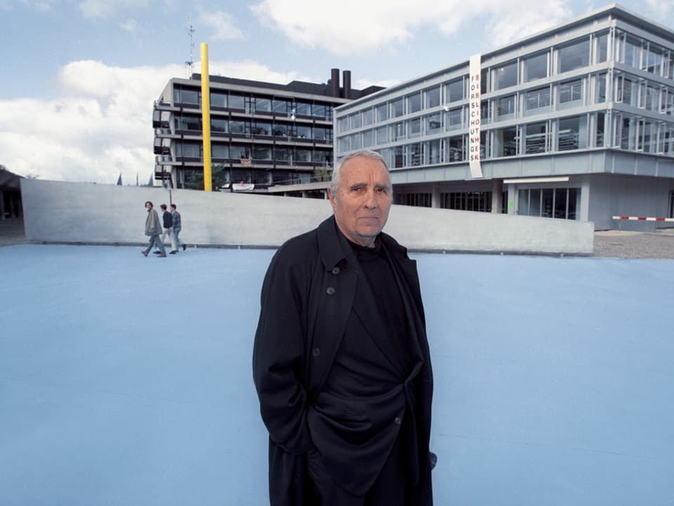 Gottfried Honegger steht auf der blauen Fläche, im Hintergrund eine Betonmauer, eine gelbe Stele und Gebäude der Universität.