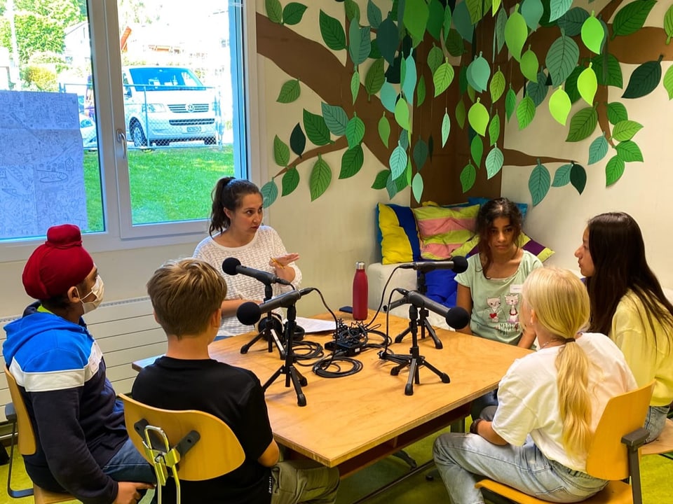 In einem Gruppenraum sitzen fünf Kinder um Mikrofone und erzählen.