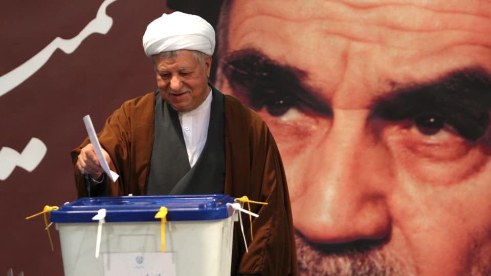 Rafsandschani, ein Mann mit weissem Turban, wirft den Stimmzettel in die Plastikurne, hinter ihm ein grosses Wandbild von Ajatollah Chomeini.