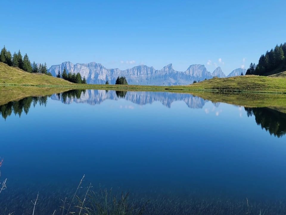 Blick auf Bergsee. Darin gespiegelt die Landschaft. Im Hintergrund sieht man Berge.
