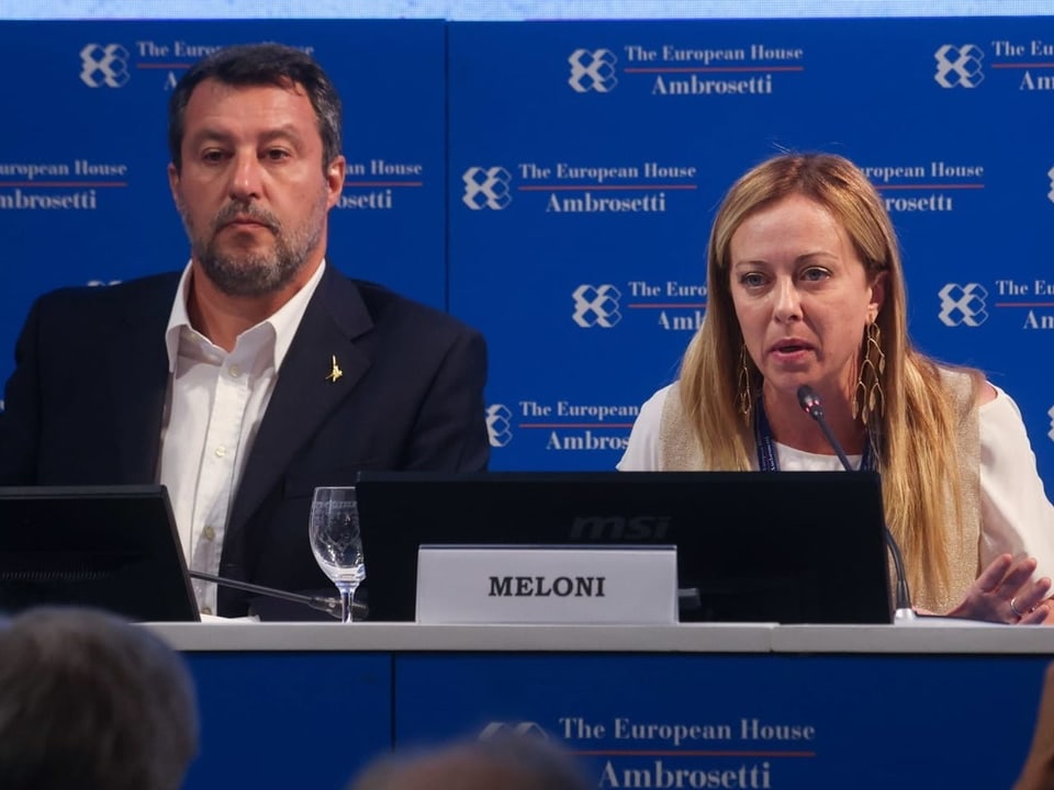 Matteo Salvini sitzt (links) neben Giorgia Meloni auf einem Podium.