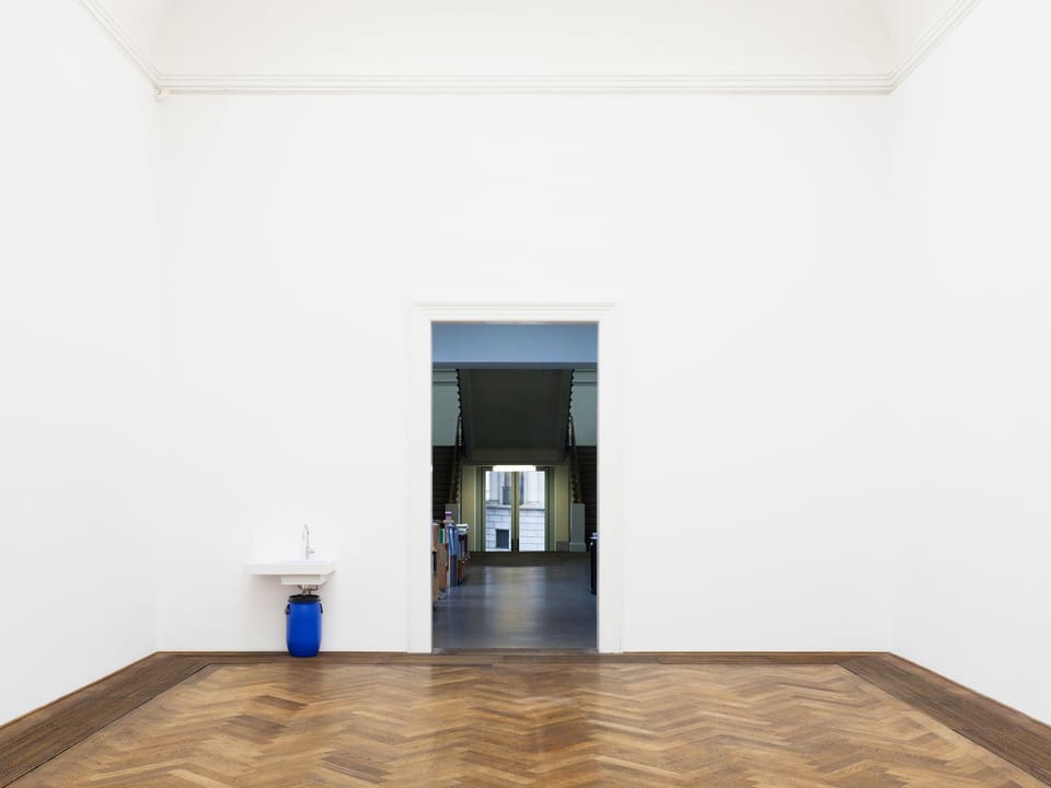 Eine Installation aus einem blauen Eimer und einem Lavabo in einem hohen weissen Raum mit Parkett.