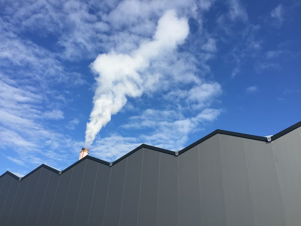 Blick an eine graue Wand einer grossen Lagerhalle mit einem rauchenden Kamin auf dem Dach.