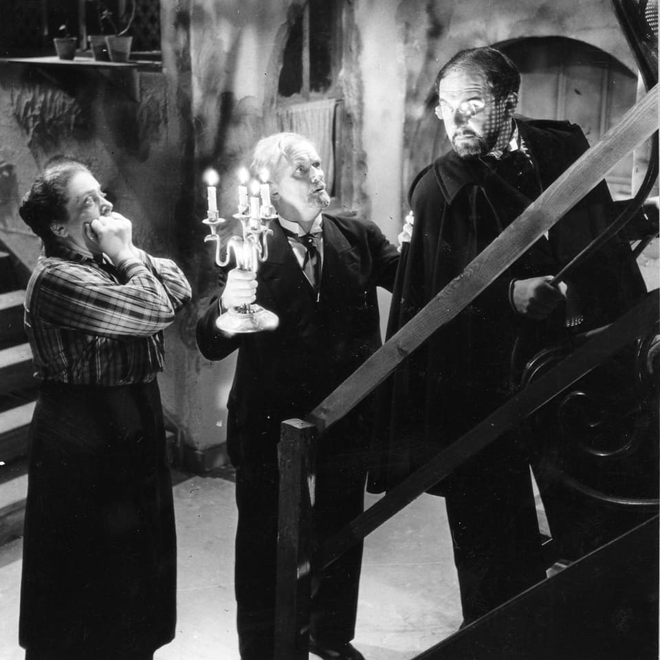 Zwei Männer und eine Frau stehen in einem Keller. Der Mann in der Mitte hält einen Kerzenhalter.