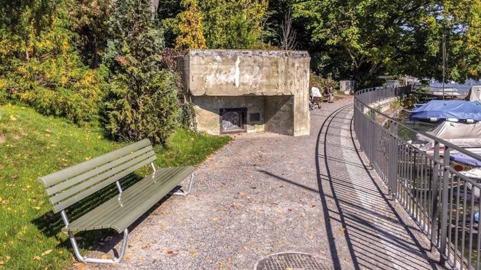 Ein Bunker an der Seepromenade, im Vordergrund ein Kiesweg, links eine SItzbank, dahinter der Bunker und Bäume.