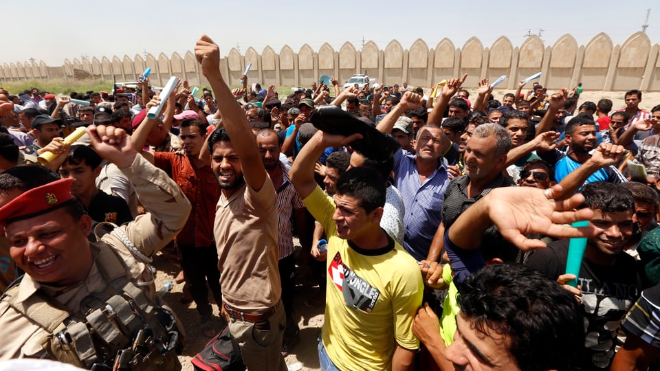 Männer in Reih und Glied skandieren Parolen, einige haben die Arme erhoben, zu sehen auch ein irakischer Soldat.