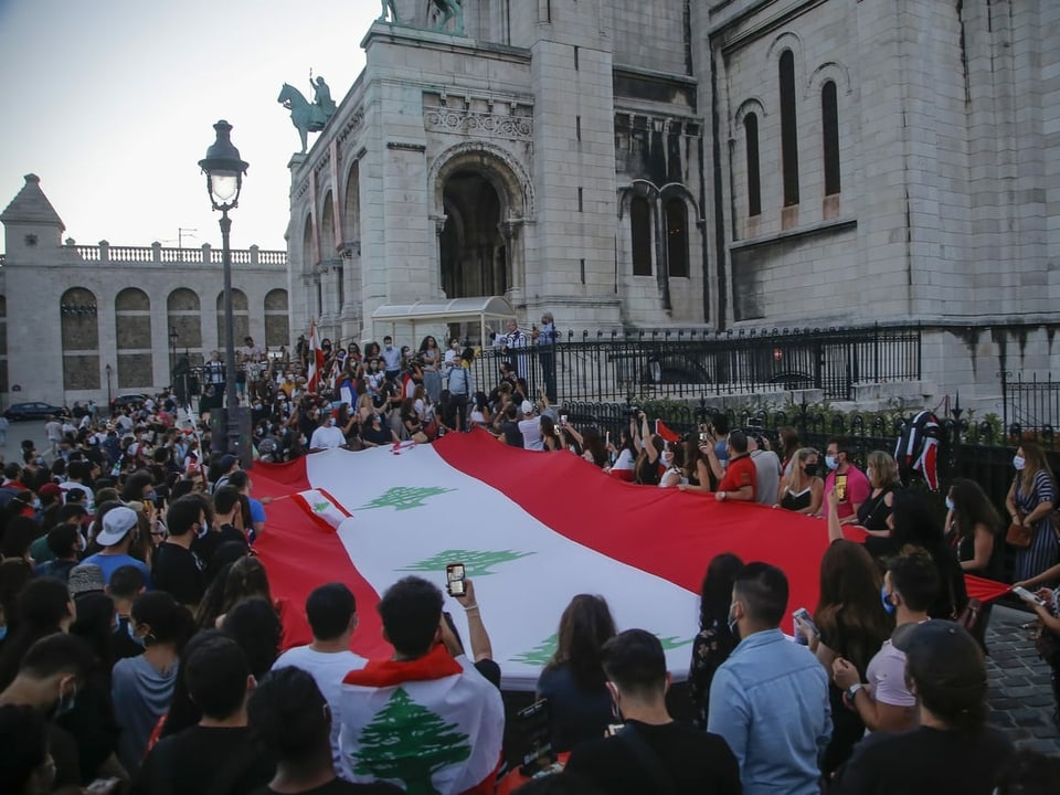 Viele Menschen stehen vor einer Kathedrale und halten eine grosse Libanon-Flagge hoch.