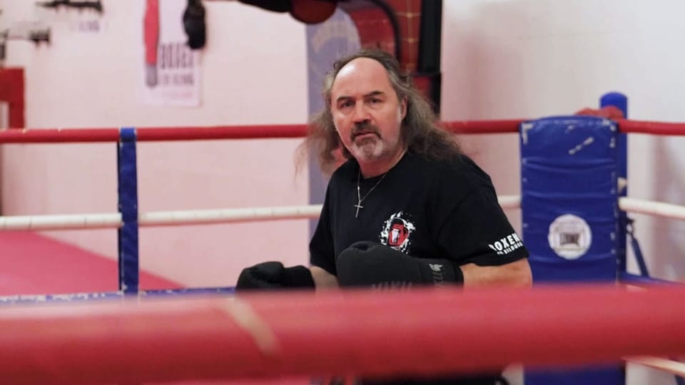 älterer Mann im Boxring, Gesicht zur Kamera, längere Haare, schwarze Boxhandschuhe.