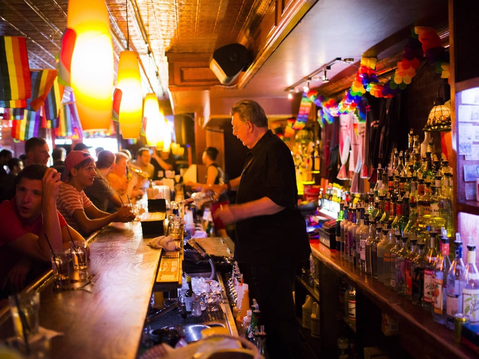 Eine Bar, man sieht den Barkeeper, ein älterer Herr mit grauen Haaren und Brille beim Ausschenken. Überall hangen Flaggen in Regenbogenfarben. Die Bar ist gut besucht.