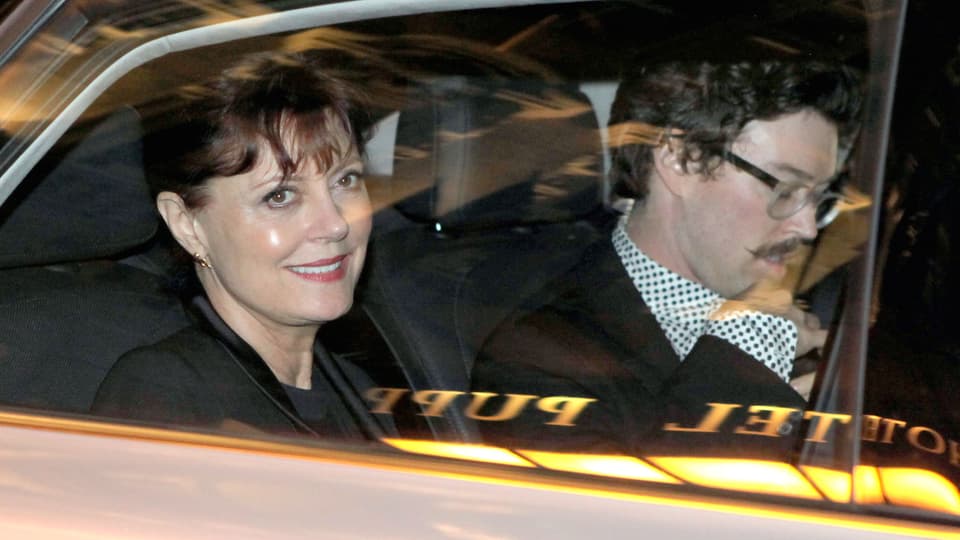 Susan Sarandon sitzt im Auto und lächelt aus dem Fenster. Daneben sitzt ein Mann mit gesenktem Blick, Brille und Schnauz.
