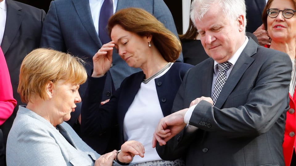 Merkel und Seehofer stehen beieinander, beide schauen auf ihre Uhren, auch andere Leute stehen drumherum.